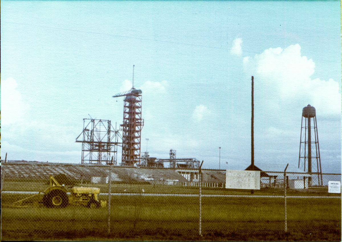 Image 004. Viewed from the turn in the road where the pad access road from A1A meets the pad perimeter, just outside the perimeter fence, the growing steel skeleton of the RSS stands in sharp relief against the sky, supported by the temporary falsework framing upon which it is resting. In this generalized view of Space Shuttle Launch Pad 39-B at the Kennedy Space Center, Florida, you can see the fifty-foot high concrete mass of the pad itself, and more or less from left to right, the RSS upon its falsework, the tall red multi-platformed framing of the FSS, the 9099 Building, the MLP Access Stair Tower, the North Piping Bridge, with the High Pressure Gas Area cutout into the pad slope beneath it, and beneath that, the supports for the cross-country piping run leading to the Hypergol Oxidizer (N2O4) Storage Facility which is partially-obscured by signage on the pad perimeter fence. At far right stands the SSW Water Tower. Photo by James MacLaren.